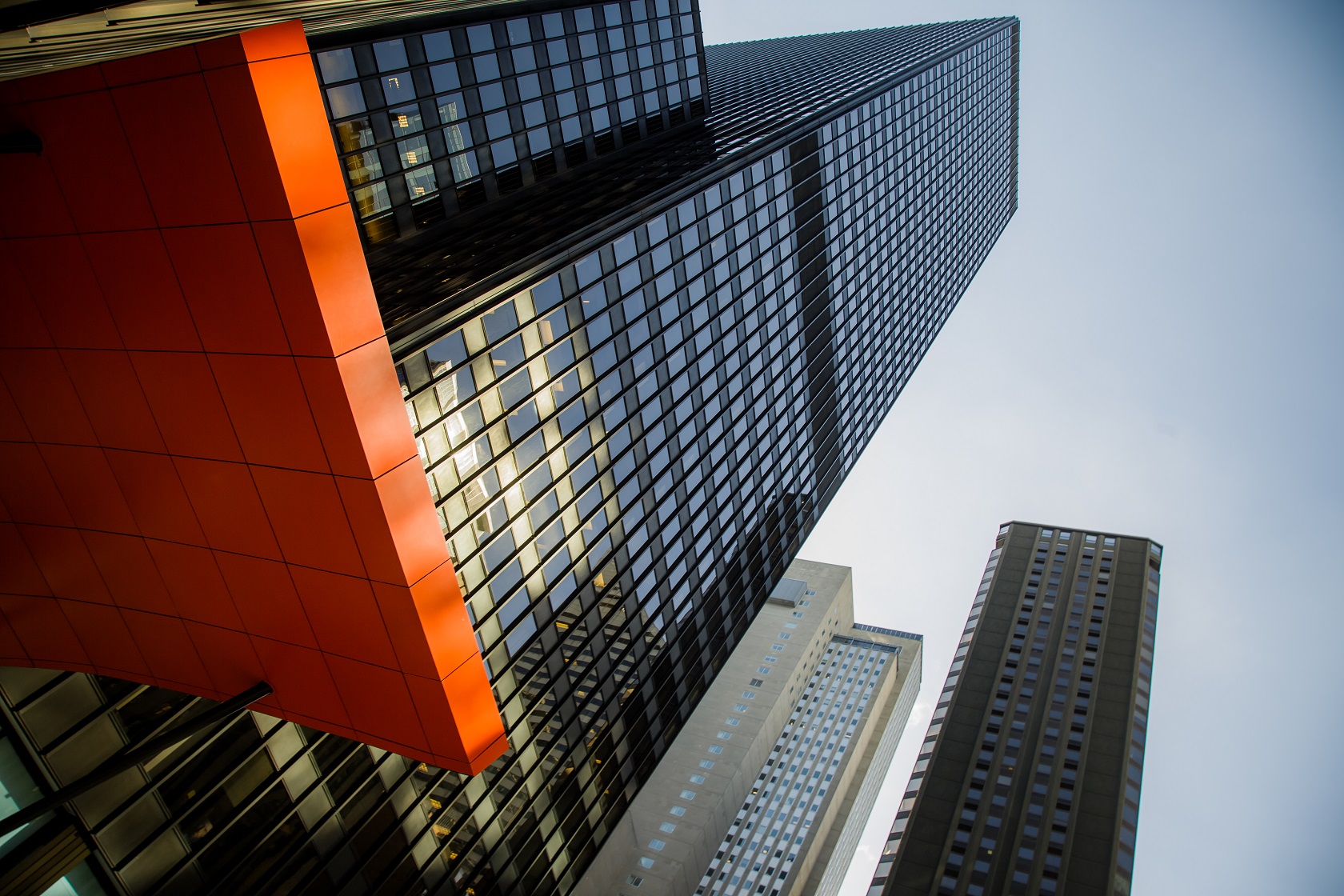 Upward view of modern skyscrapers with red-orange accent panel and gridded glass facades against blue sky.