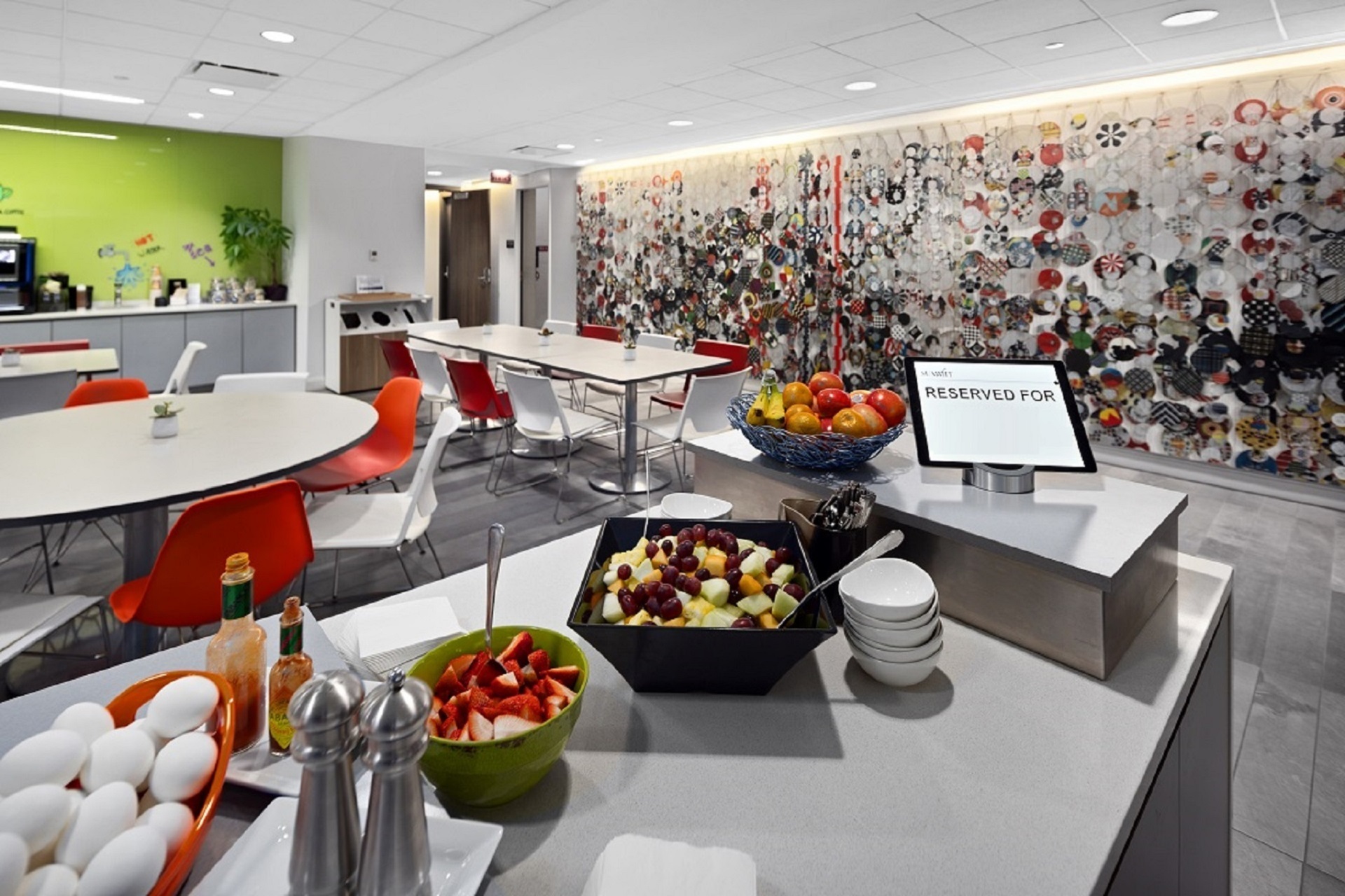 Modern office break room with breakfast spread, colorful furniture, and artistic wall collage.